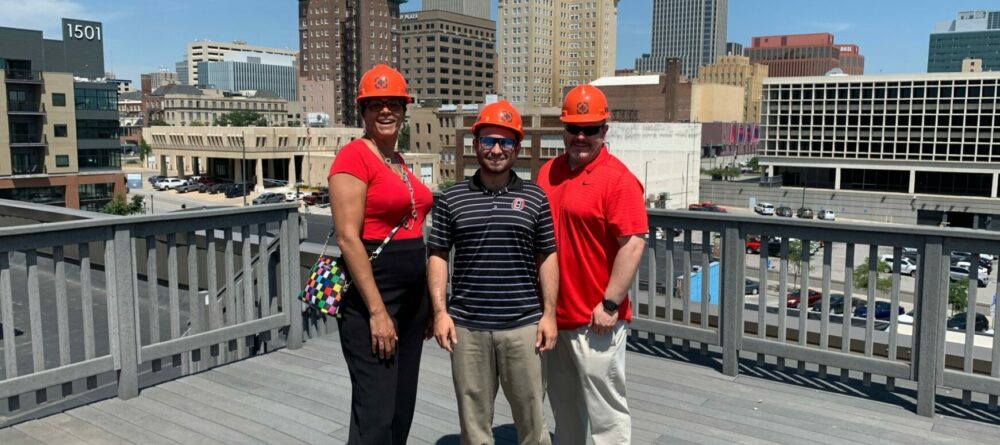 Three SourceLink Nebraska team members standing with hardhat and the Omaha skyline in the background.