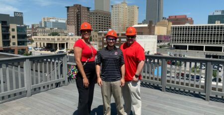 Three SourceLink Nebraska team members standing with hardhat and the Omaha skyline in the background.